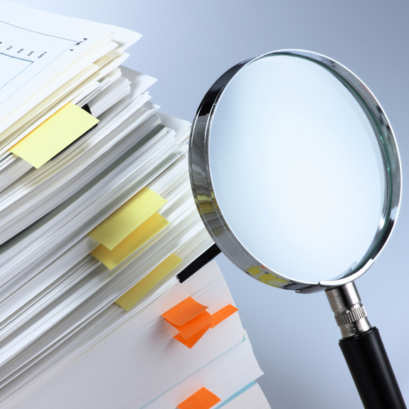 Image of a Large Stack of Papers with a Magnifying Glass Pointed at the Stack